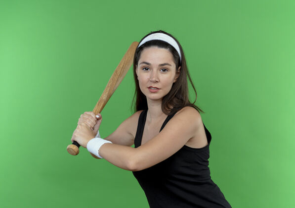 女子戴着头巾 挥舞着棒球棒 自信地站在绿色墙壁上的年轻健身女士健身棒球站立