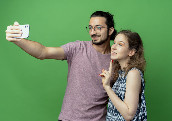 拍摄年轻漂亮的一对穿着休闲服的男女 快乐的男人用智能手机拍下他们站在绿色的墙上照片衣服男人
