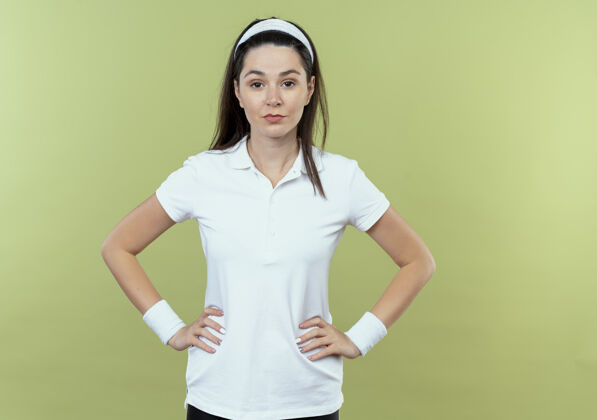 女人戴着头巾 表情自信的年轻健身女士 双臂放在臀部 站在轻盈的墙壁上手臂头带表情