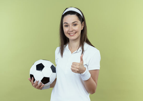 微笑戴着头巾 微笑着拿着足球 竖起大拇指站在轻质墙上的年轻健身女士举行立场足球
