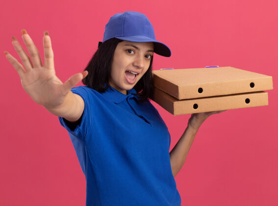 披萨情绪激动的年轻送货女孩 穿着蓝色制服 戴着帽子 拿着披萨盒 张开手掌站在粉红色的墙上 做着停下来的手势制服打开女孩