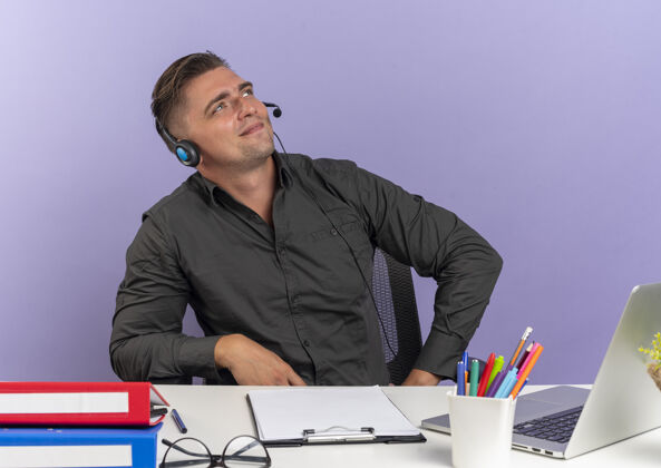 笔记本电脑年轻而快乐的金发上班族戴着耳机坐在办公桌旁 拿着办公工具 用笔记本电脑 在紫罗兰色的空间和复印空间里孤立地抬头看看请办公桌