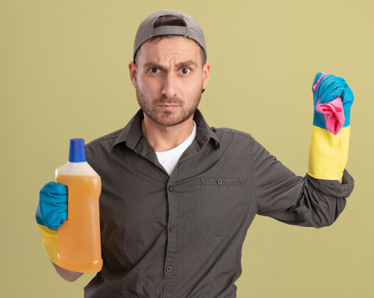 男人年轻的清洁工穿着休闲服 戴着橡胶手套 手里拿着装有清洁用品的瓶子和抹布 站在绿色的墙上怒气冲冲地看着抹布年轻瓶子