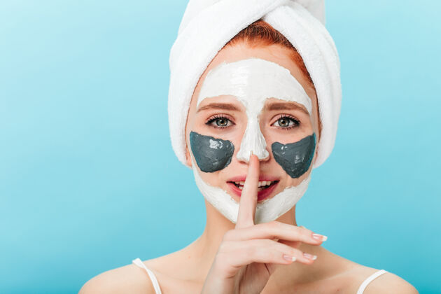治疗戴着面具的令人毛骨悚然的女士的正面图显示了沉默的标志放松的女模特头上戴着毛巾在蓝色背景下的摄影棚拍摄皮肤健康清洁面部