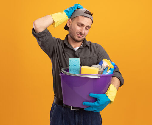 头年轻人穿着休闲服 戴着橡胶手套戴着帽子 手里拿着水桶 手里拿着清洁工具 站在橘色的墙上 头上放着一只手 看上去既困惑又不高兴手套男人立场