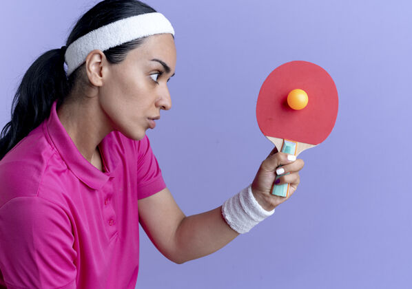 复制年轻自信的白人运动女性戴着头带和腕带 拿着乒乓球拍和球 在紫色空间与复制空间隔离球球拍举行
