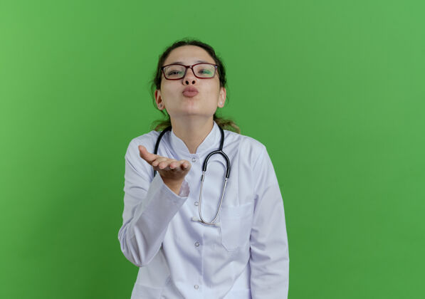 医生自信的年轻女医生穿着医用长袍 戴着听诊器 戴着眼镜 送着口吻 把手放在空气中 隔离在绿色的墙上 留着复印空间看保持医疗