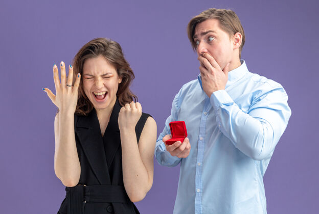 手指年轻漂亮的夫妇困惑和震惊的男子与红盒子看着他兴奋的疯狂快乐的女友与订婚戒指手指情人节的概念站在蓝色背景男人女朋友概念