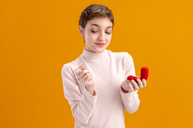 年轻一位留着短发的年轻女子手持红色盒子和订婚戒指面带微笑地看着它站在橙色的墙上情人节的概念脸订婚微笑