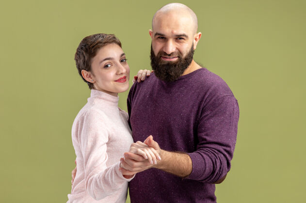 站一对穿着休闲服的年轻夫妇一个留着短头发和胡须的女人在爱中快乐地跳舞庆祝情人节站在绿色的墙上男人庆祝年轻