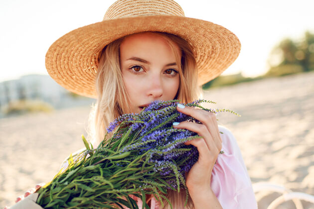 健康戴着草帽的温柔漂亮的女人在海边阳光明媚的海滩上摆着一束鲜花特写肖像头发眼睛衣服