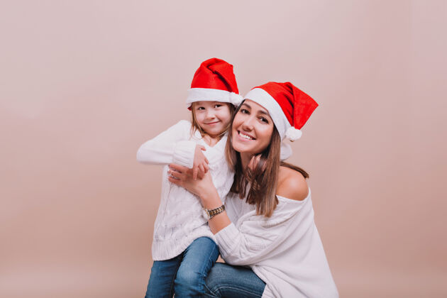 快乐美丽的母亲和小可爱的女儿穿着白色套头衫和圣诞老人的帽子画像节日爱可爱