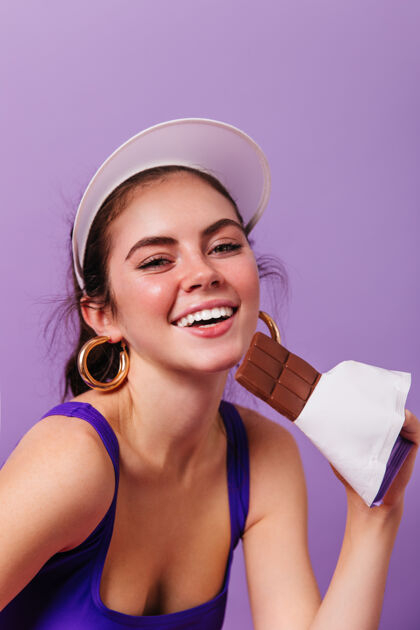 魅力戴着亮金耳环 戴着白帽子的年轻女子微笑着 手里拿着一块巧克力放在紫色墙上的特写肖像漂亮皮肤美丽