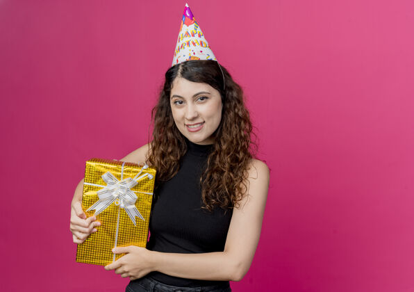 女性一位年轻漂亮的女士 卷发 戴着节日帽 手里拿着生日礼盒 脸上洋溢着欢笑 生日派对的概念是粉色的头发礼物微笑