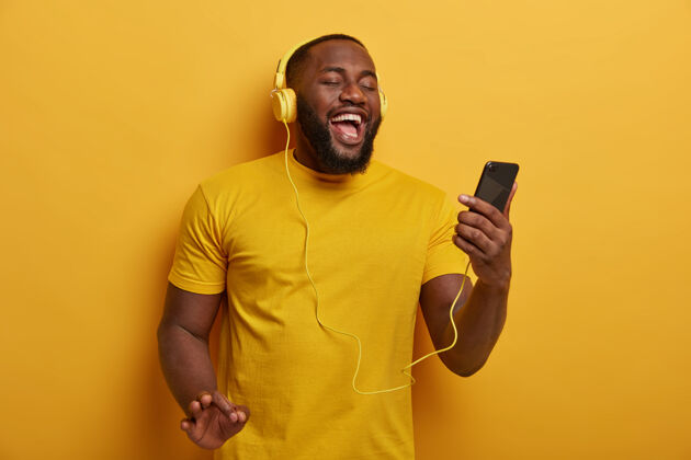 快乐半身镜头的黑人听音乐冷 持有现代智能手机 戴着耳机的耳朵 享受美好的轨道 姿势在黄色的背景微笑设备男人