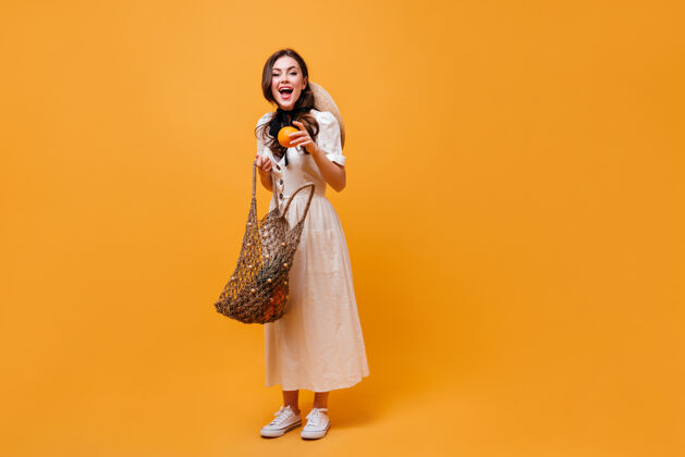 包快乐的女人从线袋子里拿出橙色穿着白色迷笛裙的女士在橙色背景上摆姿势女士水果连衣裙
