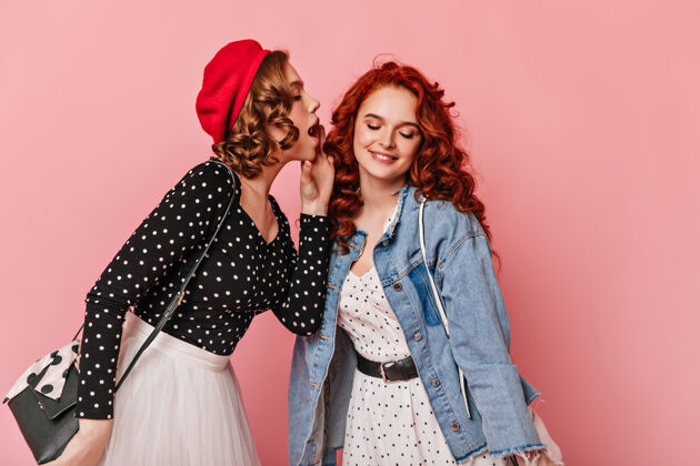 手提包在粉色背景下分享秘密的迷人女孩穿着时髦衣服的会说话的女士的摄影棚照片贝雷帽红发优雅