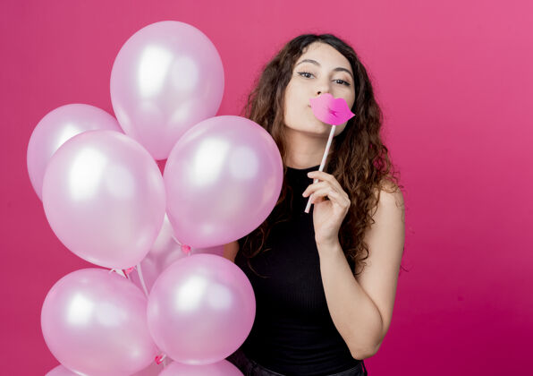 抱着一个年轻漂亮的女人 卷发 手里拿着一束气球 手里拿着派对用的棍子 站在粉红色的墙上开心地笑着年轻气球欢呼