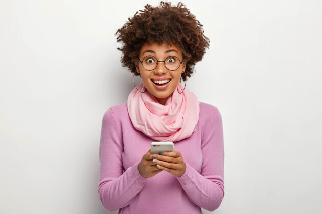 眼镜快乐的卷发女人手持现代手机 在线聊天 开心地微笑 戴着透明眼镜和休闲的紫色套头衫站着情绪媒体