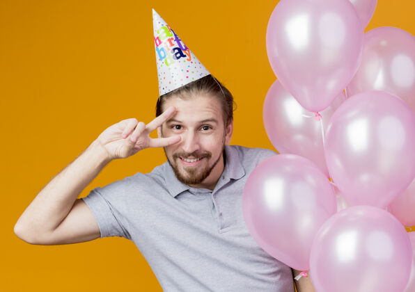 庆祝戴着节日帽的年轻人举着一堆气球欢笑着庆祝生日 橙色的墙上挂着v字标志气球立场举行