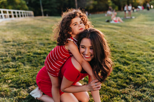 孩子一头卷发的放松的孩子拥抱着大自然中的母亲穿着红色连衣裙的惊艳女人和女儿在公园里嬉戏年轻拥抱快乐