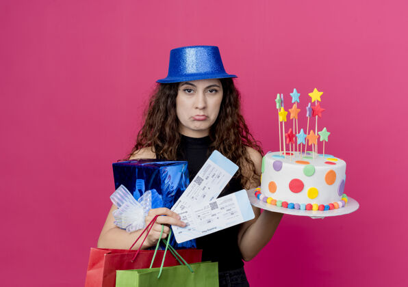 机票一位年轻漂亮的女士 卷发 戴着节日礼帽 手里拿着生日蛋糕礼盒和机票 脸上带着悲伤的表情 站在粉色的墙上 生日派对的概念看着蛋糕表情