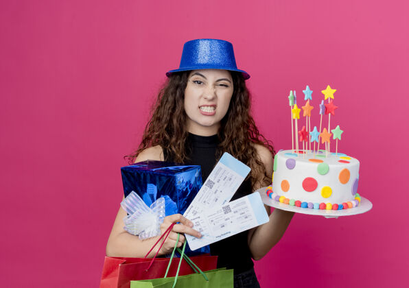 蛋糕一个年轻漂亮的女人 卷发 戴着节日礼帽 手里拿着生日蛋糕礼盒和机票 站在粉色的墙上 看起来困惑和不高兴的生日派对概念年轻看着卷曲