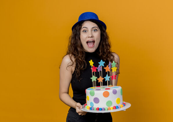 兴奋一位年轻漂亮的卷发女士 戴着节日礼帽 手里拿着生日蛋糕 站在橘色的墙上 快乐而兴奋卷发帽子年轻