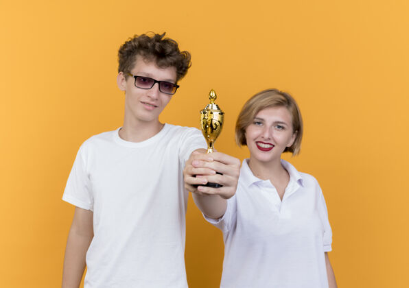 表演年轻的运动情侣男女站在一起展示奖杯微笑着幸福的脸站在橙色的墙上脸奖杯在一起