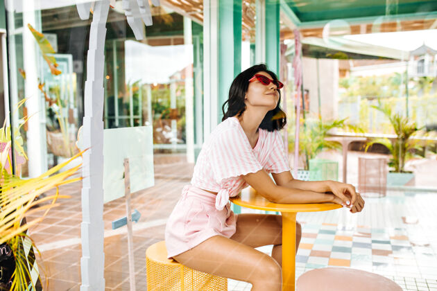 晒黑穿着短裤微笑的女人坐在户外咖啡馆里戴着粉色太阳镜的梦幻女人享受着夏日周末生活方式黑发欧洲