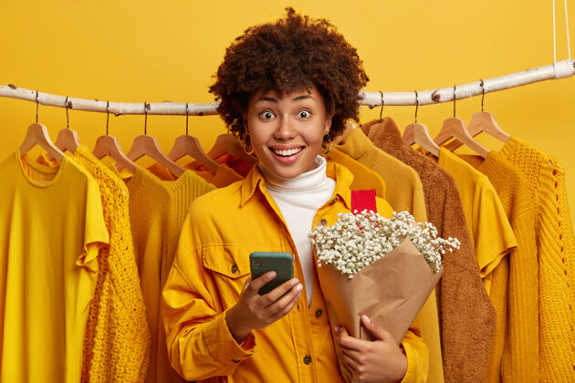 电话笑容可掬的美国黑人女士手持花束和现代手机 在挂在背景上的衣架旁摆姿势明亮高兴商店