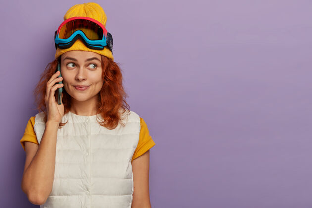外套滑雪场上可爱的年轻姜女横拍 打电话 戴着黄帽子和白背心 站在室内紫色的墙上高兴白种人运动