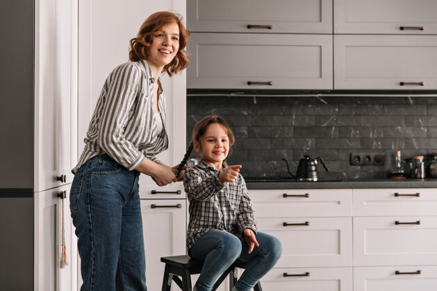 牛仔裤穿着衬衫和牛仔裤的年轻母亲站在坐在椅子上的女儿旁边食物欢呼房子