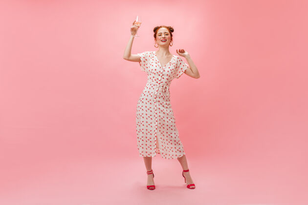 简介迷人的女人穿着时髦的白色圆点裙 在粉色背景上与一杯香槟翩翩起舞美女微笑欢呼
