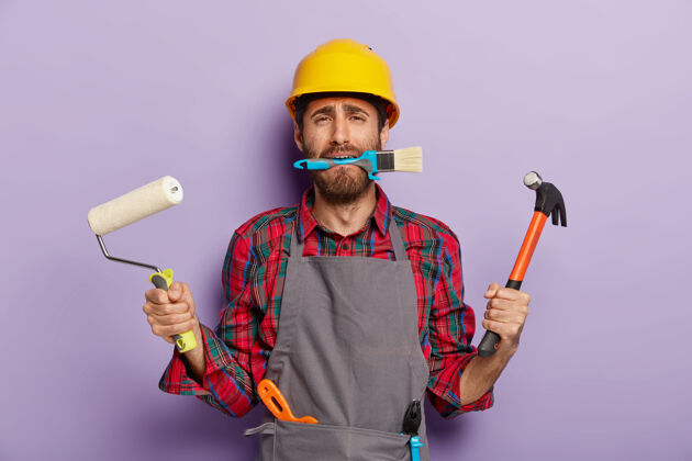 男忙碌的修理工拿着建筑工具 在家里修理 戴着黄色的安全帽 围裙 站在室内工具工作不高兴