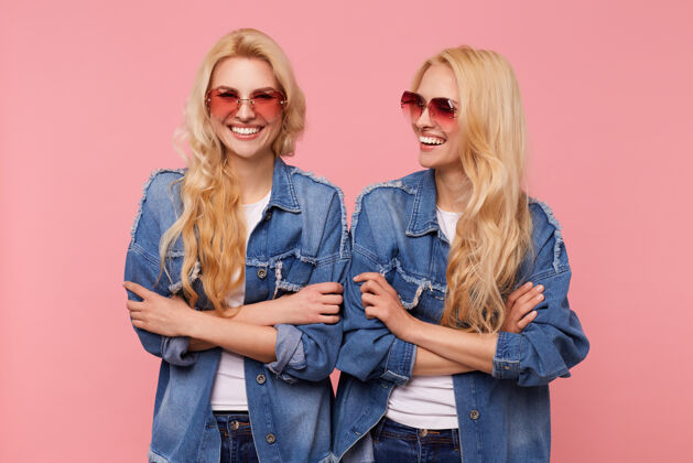 金发快乐的年轻长发金发女性 戴着太阳镜 穿着休闲服 双手合十 在粉色背景上摆姿势 开心地笑着头发双胞胎休闲