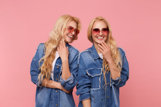 双胞胎室内拍摄的年轻可爱的快乐的长发金发双胞胎穿着同样的衣服在一起玩得很开心 开心地笑 而站在粉红色的背景Swoosh牛仔裤白皙