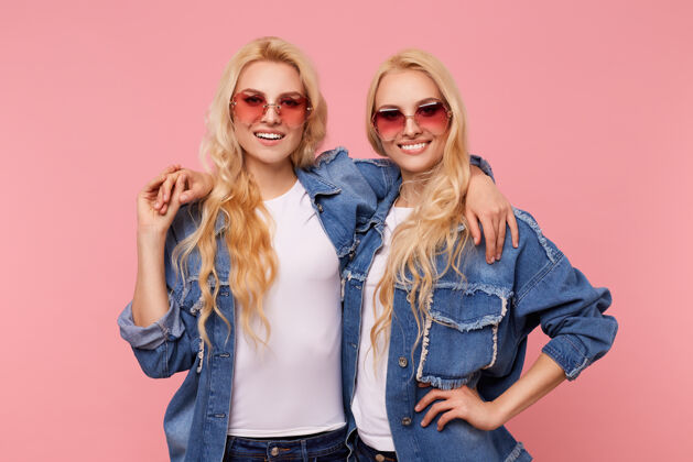 高兴迷人的年轻快乐的长发白发姐妹戴着红色太阳镜 站在粉色的背景下 深情地拥抱着对方 开心地看着镜头红色太阳镜拥抱