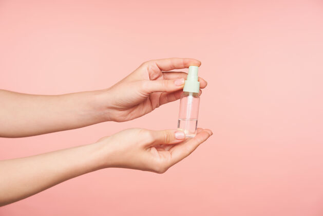 皮肤白皙室内拍摄的裸体美甲举起女性的手拿着消毒喷雾瓶 同时被隔离在粉红色背景上人类的手的概念成人透明女性