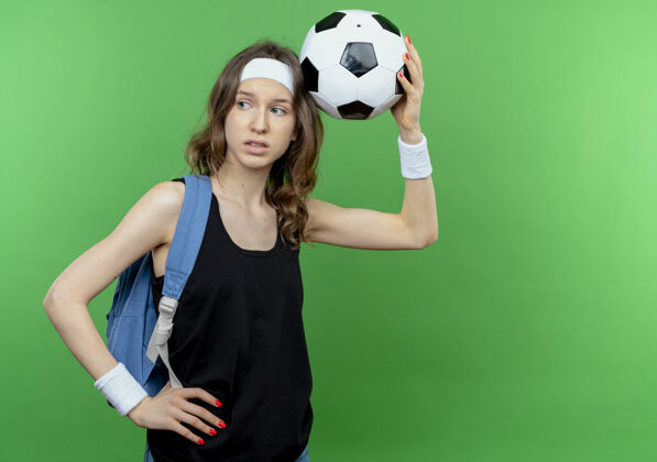 健身穿着黑色运动服 背着背包 头箍 头上举着足球的年轻健身女孩站在绿色的墙上 一边困惑地看着拼图立场女孩