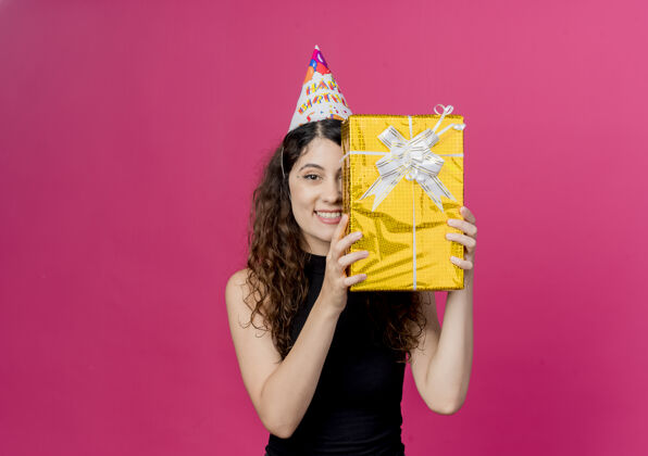 年轻一位年轻漂亮的卷发女士 戴着节日帽 手持生日礼盒 微笑着 站在粉色的墙上 高高兴兴地庆祝生日女人欢呼卷发