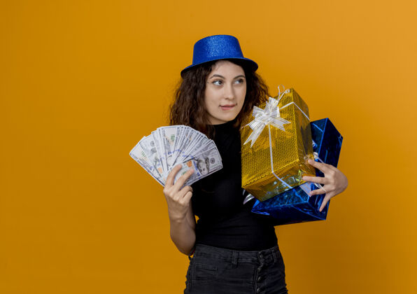 欢呼年轻漂亮的女人 卷发 戴着礼帽 手里拿着现金和礼物 看着卡内拉快乐地站在橙色的墙上看着年轻现金