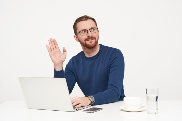 套头衫帅气的年轻人 一头金发 身穿蓝色毛衣 抬起手掌打招呼 一边看一边 在白色的背景下用笔记本电脑工作杯子男性蓝色