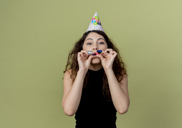 年轻年轻漂亮的女人卷发在节日帽子吹哨子快乐和积极的生日派对概念站在轻墙哨子吹站