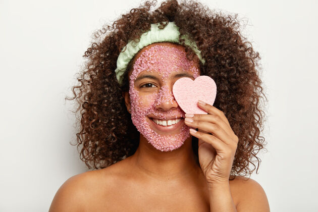 卫生可爱可爱的卷曲非洲女子的水平镜头微笑 涂抹去皮面膜 用海绵覆盖眼睛 呵护皮肤 周末在家有美容套路 健康的样子舒缓化妆品裸露
