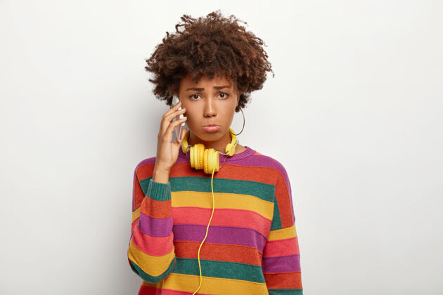 条纹闷闷不乐的非裔美国女孩通过智能手机进行不愉快的交谈 穿着色彩鲜艳的休闲条纹套头衫 使用黄色耳机 对某事不满闷闷不乐女人美国人