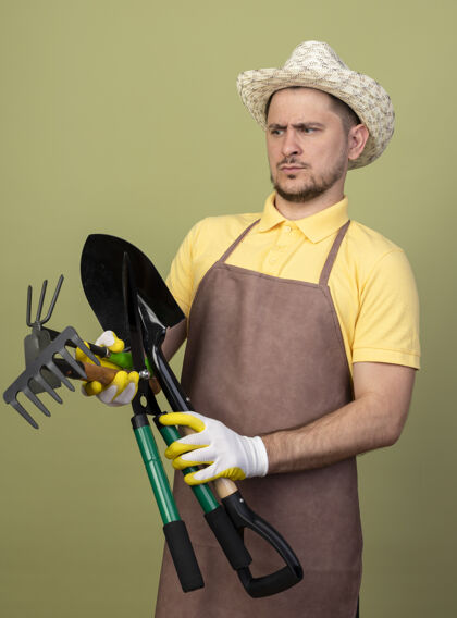 抱年轻的园丁穿着连体衣戴着帽子戴着工作手套拿着园艺设备看着他们不高兴帽子连身衣男人