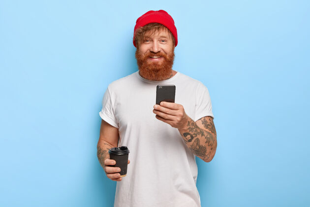 男人半身镜头：快乐的红胡子男人戴着时髦的帽子 穿着白色休闲t恤 拿着手机 喝着外卖咖啡 心情很好 打字留言电话纹身在线