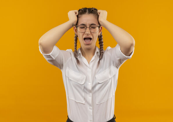 头发一个戴着眼镜 梳着辫子 穿着白衬衫的沮丧的年轻女学生站在橙色的背景上 拉着头发发狂野性橙色辫子