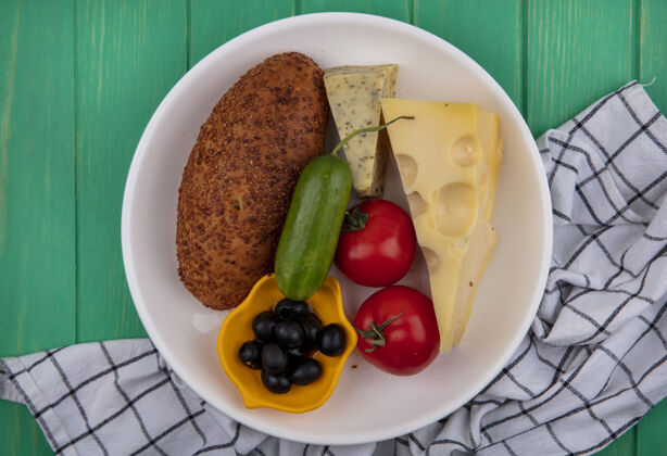 橄榄白色盘子上芝麻饼的俯视图 绿色木制背景上格子布上有新鲜蔬菜 奶酪和橄榄肉饼肉食物